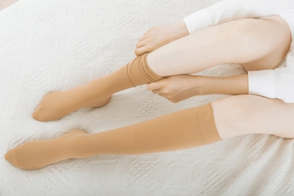 An individual is shown in a white shirt, sitting on a bed and putting on a compression sock.