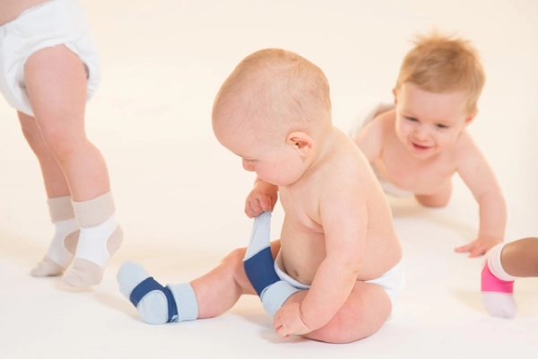 Three adorable babies are wearing infant socks, with one sitting down and pulling on a blue sock.