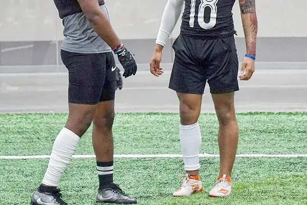 Two football players on a field, one wearing a white one leg sleeve football sock.